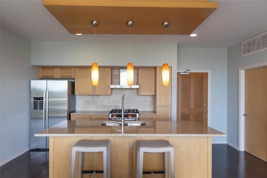 Kitchen with backsplash, stainless steel fridge, light stone countertops, pendant lighting, and a center island with sink