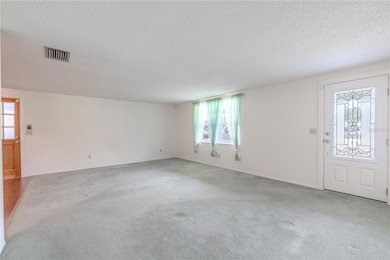 This spacious family room offers a clean and versatile canvas, ready to accommodate a variety of furniture layouts and design styles. A large front-facing window fills the room with natural light, while the neutral wall color and soft carpet flooring enhance the sense of openness. The decorative glass-panel front door adds architectural interest and additional brightness to the space.