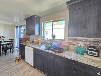 Kitchen with dark brown cabinets, white dishwasher, light wood-type flooring, and light stone countertops