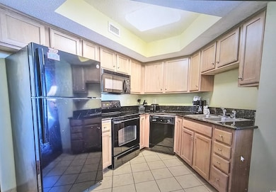 Kitchen featuring black appliances, a tray ceiling, light tile patterned floors, dark stone countertops, and light brown cabinetry