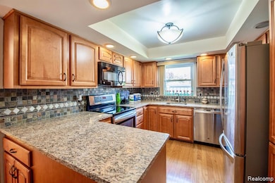 Kitchen featuring brown cabinets, stainless steel appliances, a tray ceiling, light stone counters, and decorative backsplash