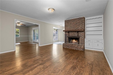 Unfurnished living room featuring a textured ceiling, a brick fireplace, dark wood finished floors, built in shelves, and a ceiling fan