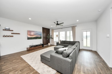 This modern living room offers a spacious, open layout designed for both comfort and style. Natural light pours in through large windows and a glass door, highlighting the rich hardwood flooring and contemporary finishes. A large sectional sofa invites relaxation, while a mounted TV, sleek built-in shelving, and a ceiling fan add function and flair to this inviting space—ideal for everyday living and entertaining alike.

