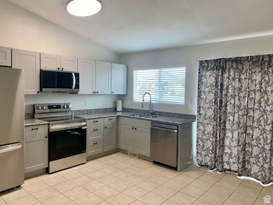 Kitchen featuring stainless steel appliances, vaulted ceiling, gray cabinetry, dark stone counters, and light tile patterned flooring