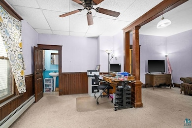 Office area with a baseboard radiator, a paneled ceiling, carpet, a wainscoted wall, and ceiling fan
