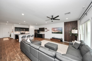 This photo highlights a beautifully designed open-concept living space that seamlessly blends a sleek, modern kitchen with a spacious living area. Warm wood flooring, a comfortable sectional sofa, and contemporary touches like recessed lighting and a ceiling fan create a stylish yet inviting atmosphere. Expansive windows flood the space with natural light, making it perfect for both everyday living and entertaining.