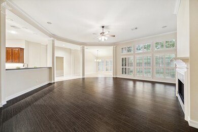 This is a spacious, well-lit room with elegant dark hardwood flooring, crisp white trim, built-in shelving, and plantation shutters on the windows. The room features a modern chandelier and has a neutral color palette, offering a blank canvas for personalization.