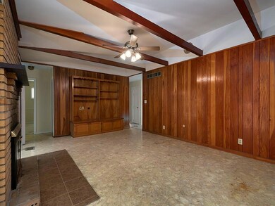 Unfurnished living room featuring wood walls, beamed ceiling, ceiling fan, and a brick fireplace