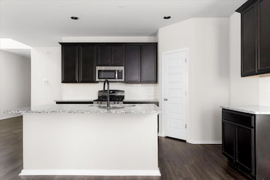 Kitchen featuring decorative backsplash, light stone counters, appliances with stainless steel finishes, dark wood-type flooring, and a center island with sink