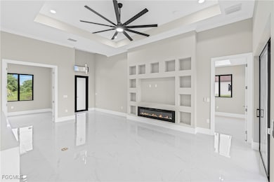 Unfurnished living room with built in features, light marble finish floors, a raised ceiling, ceiling fan, and ornamental molding