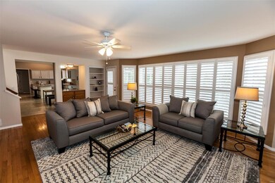Living room with hardwood flooring, ceiling fan with light, bay window & built in bookshelves.