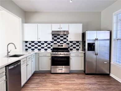 Kitchen with appliances with stainless steel finishes, white cabinetry, under cabinet range hood, light stone counters, and dark wood-style floors