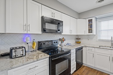 Kitchen with black appliances, tasteful backsplash, white cabinets, glass insert cabinets, and light stone countertops