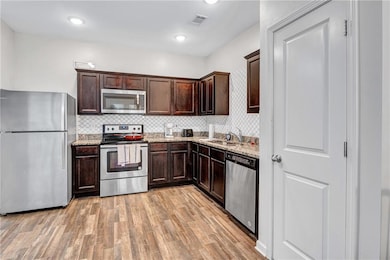 Kitchen featuring appliances with stainless steel finishes, dark brown cabinetry, decorative backsplash, light wood-type flooring, and light stone countertops