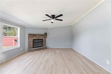 Unfurnished living room with light wood-type flooring, a textured ceiling, a stone fireplace, a ceiling fan, and ornamental molding
