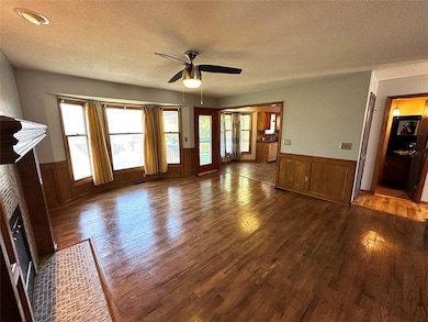 Unfurnished living room with a wainscoted wall, dark wood-style flooring, a textured ceiling, ceiling fan, and a fireplace with flush hearth