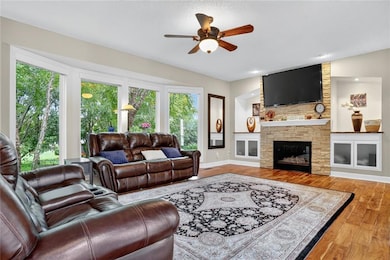 Living area with wood finished floors, a fireplace, and ceiling fan and 4 very large windows with a view of mature trees