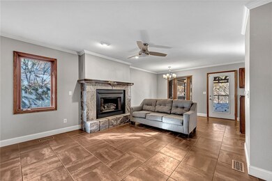 Tiled living room featuring crown molding, a stone fireplace, a chandelier, and ceiling fan