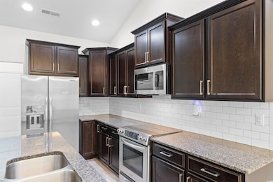 Kitchen with appliances with stainless steel finishes, light stone countertops, dark brown cabinets, vaulted ceiling, and backsplash
