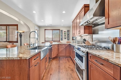 Kitchen featuring appliances with stainless steel finishes, light stone counters, brown cabinetry, wall chimney exhaust hood, and recessed lighting
