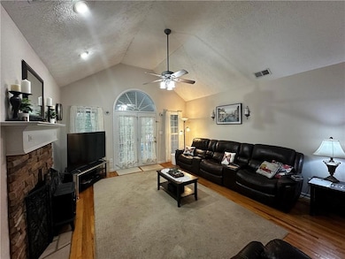 Living room with a textured ceiling, a stone fireplace, wood finished floors, a ceiling fan, and high vaulted ceiling