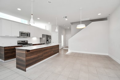 Modern kitchen with white cabinetry, stainless steel appliances, an island with dark wood finish, and pendant lighting, adjacent to a spacious, well-lit area leading to a staircase.