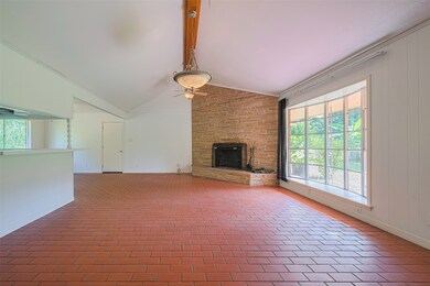 This spacious living room features a vaulted ceiling with an exposed beam, large windows for ample natural light, a cozy brick fireplace, and terracotta tile flooring, creating a warm and inviting atmosphere.