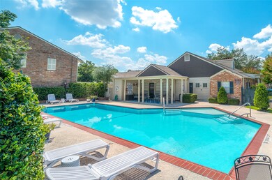 This photo showcases a beautiful community pool area with a spacious, well-maintained pool, surrounded by lounge chairs and lush greenery. Adjacent to the pool is a charming brick clubhouse, offering a relaxing and inviting atmosphere.