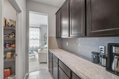 This photo showcases a stylish butler's pantry featuring rich dark wood cabinetry, quartz countertops, and a sleek subway tile backsplash. Adjacent to the space is a walk-in pantry with ample wire shelving for storage. The butler’s pantry flows seamlessly into a bright sitting area, creating a practical and elegant transition between kitchen and living spaces.