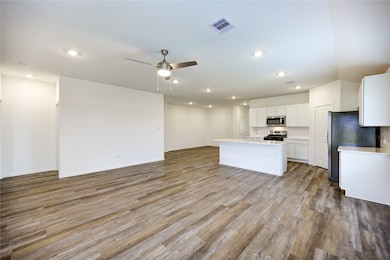 This photo showcases a spacious, open-concept living area with modern wood-look flooring. The kitchen features sleek white cabinetry, stainless steel appliances, and a large island. Recessed lighting and a ceiling fan add to the room's functionality and comfort.