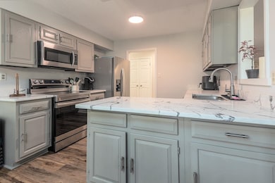 Kitchen featuring appliances with stainless steel finishes, gray cabinetry, light stone counters, dark wood finished floors, and a peninsula