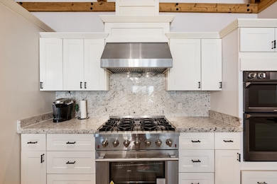 Kitchen with stainless steel stove, tasteful backsplash, under cabinet range hood, white cabinets, and multiple ovens