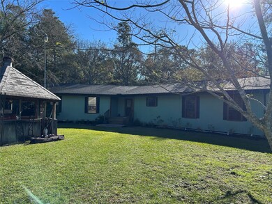 This photo shows a single-story home for rent with a spacious front yard, surrounded by mature trees. The house features a simple design with a small porch, and there's a wooden gazebo to the side. The setting is peaceful and private, perfect for enjoying nature.