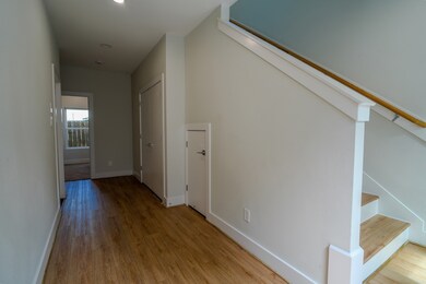 A bright and inviting entrance of a home, featuring a wooden staircase leading upstairs. Natural light streams in from a window, illuminating the light-colored walls and the polished floor.
