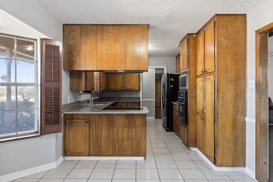 Kitchen featuring brown cabinetry, a peninsula, light tile patterned floors, light stone counters, and black appliances
