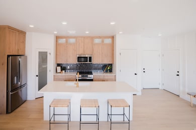Kitchen with appliances with stainless steel finishes, light brown cabinetry, tasteful backsplash, light wood-style flooring, and a kitchen island with sink