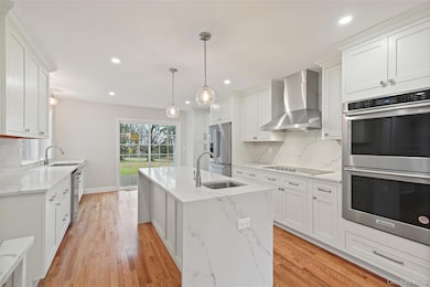 Kitchen with stainless steel appliances, tasteful backsplash, wall chimney range hood, light stone counters, and white cabinetry