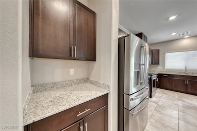 Kitchen featuring stainless steel appliances, light stone countertops, dark brown cabinetry, light tile patterned floors, and a textured wall