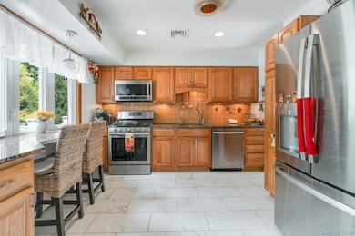 Kitchen featuring stainless steel appliances, backsplash, brown cabinetry, light marble finish floors, and dark stone counters