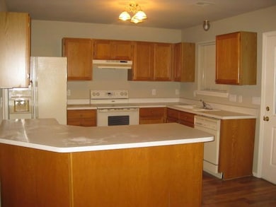 Kitchen with white appliances, under cabinet range hood, light countertops, dark wood-style floors, and brown cabinetry