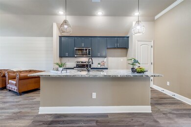 Kitchen featuring upgraded light stone countertops, appliances with stainless steel finishes, a kitchen island with sink, upgraded pendant decorative light fixtures, and wood-type flooring, upgraded custom paint and added hardware to cabinets