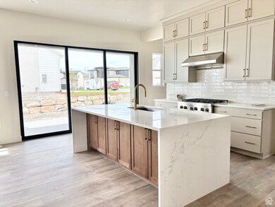 Kitchen featuring light stone counters, backsplash, an island with sink, light wood finished floors, and under cabinet range hood