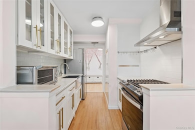 Kitchen with wall chimney exhaust hood, stainless steel gas range, decorative backsplash, light wood-type flooring, and light countertops