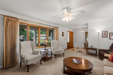 Opposite view of the living room.  Ceiling fan, front facing picture window, open staircase to the basement, and hardwood floors underneath the existing carpet.