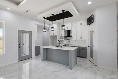 Kitchen featuring sink, white cabinetry, decorative light fixtures, an island with sink, and wall chimney range hood