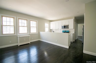 Kitchen with radiator heating unit, a peninsula, stainless steel microwave, white cabinetry, and dark wood-style floors