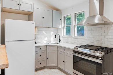 Kitchen with stainless steel gas stove, freestanding refrigerator, gray cabinetry, wall chimney range hood, and vaulted ceiling