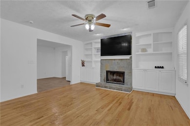 Unfurnished living room with a stone fireplace, a textured ceiling, built in shelves, light hardwood / wood-style floors, and ceiling fan