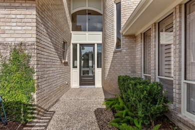 This home features a welcoming entrance with a modern, glass-paneled door surrounded by light-colored brickwork. The pathway is lined with manicured shrubs, and large windows provide ample natural light.