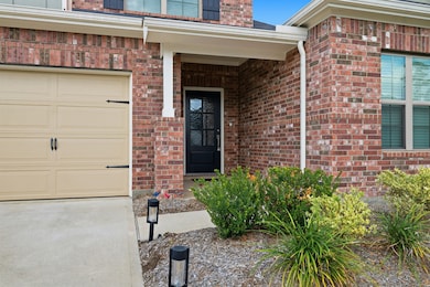 This photo shows a charming brick home exterior with a welcoming front entrance. It features a covered porch, a black front door, and a well-maintained garden. Adjacent is a beige garage door, and the area is neatly landscaped with shrubs and pathway lights.